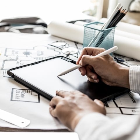 Designer sketching on tablet over printed blueprints during collaborative architectural project planning phase.